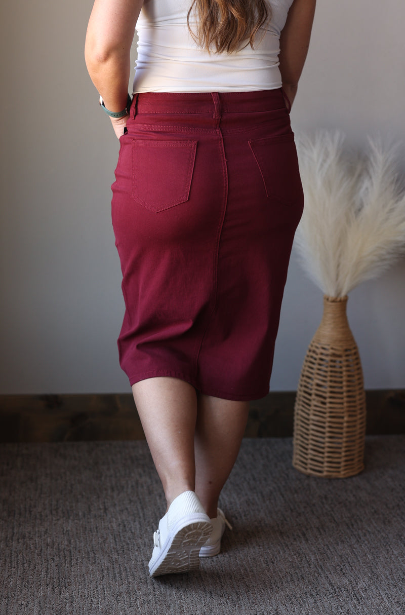 Person wearing maroon midi denim skirt with a white top and white sneakers, standing in a room with a textured wall and decorative vase.