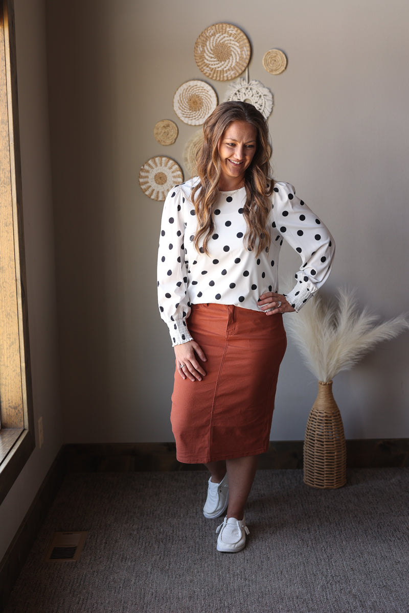 Woman wearing a polka dot blouse and rust-colored skirt standing in a room with decorative wall art.