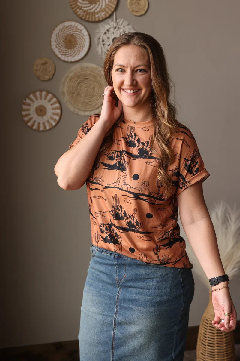 Woman wearing a patterned orange aztec shirt and blue skirt standing in a room with decorative wall art.