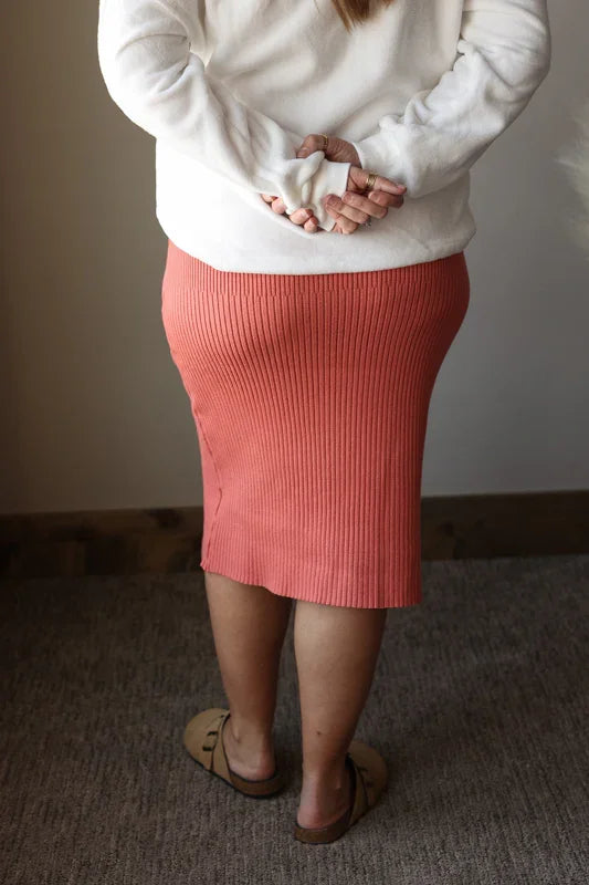 Person wearing a white sweater and pink skirt standing against a plain wall.