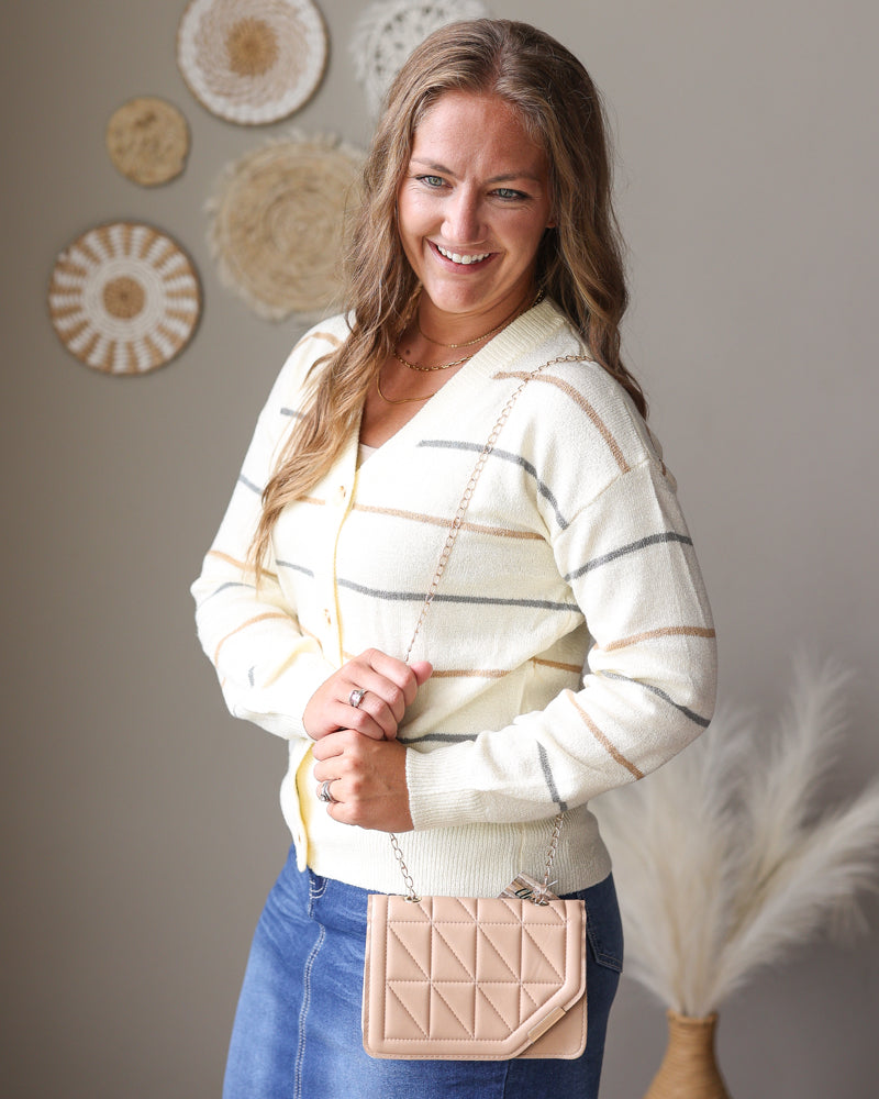A woman wearing a cream and gray striped knit cardigan sweater and a blue denim skirt, holding a beige quilted crossbody bag. She is standing in front of a neutral gray wall decorated with circular woven wall art.
