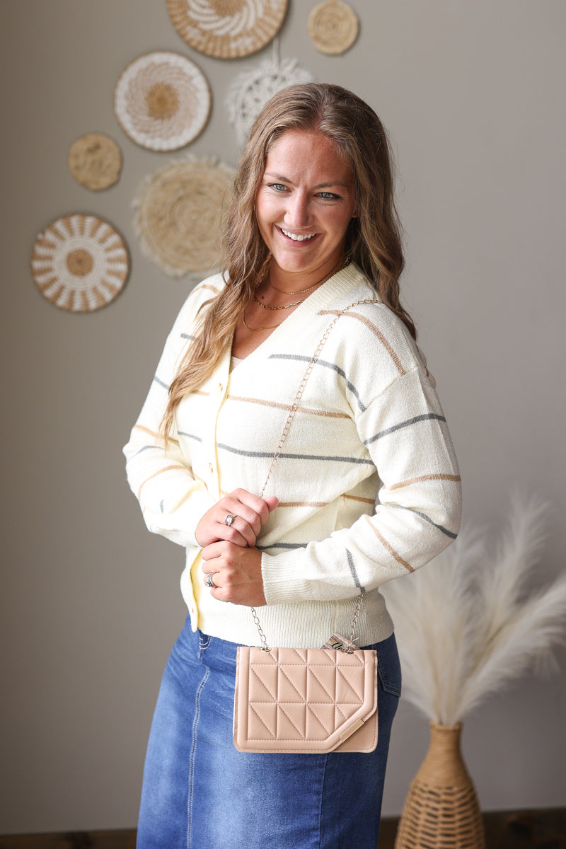 A woman wearing a cream and gray striped knit cardigan sweater and a blue denim skirt, holding a beige quilted crossbody bag. She is standing in front of a neutral gray wall decorated with circular woven wall art.