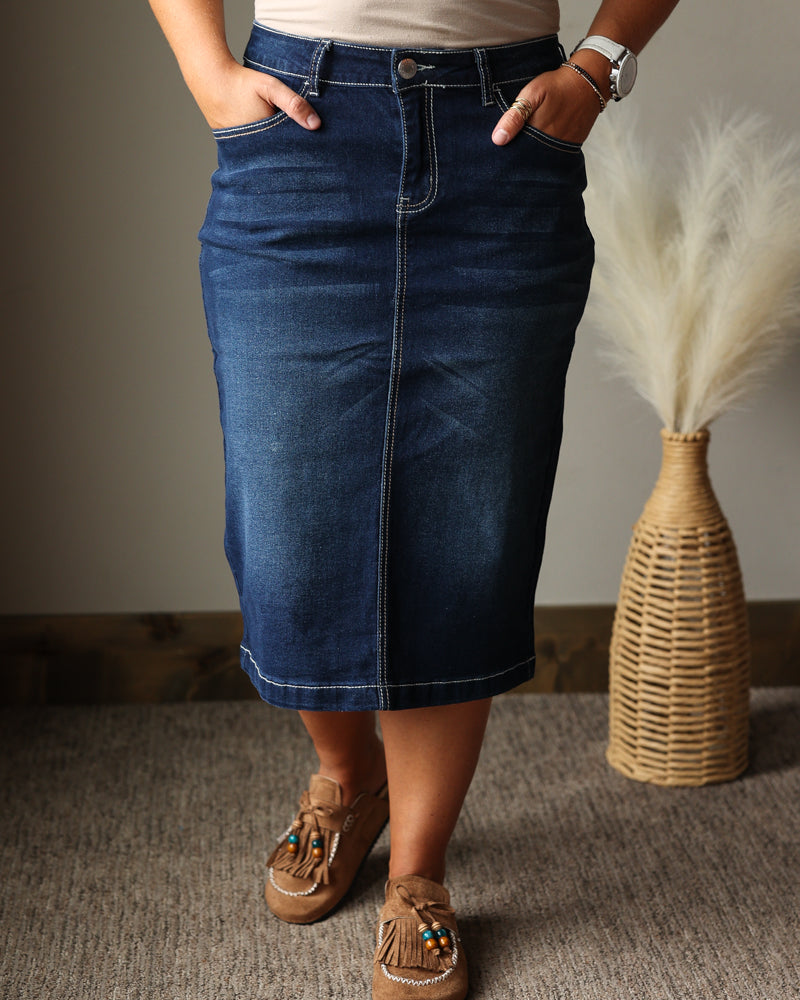 A person wearing a dark blue denim midi skirt with a straight, slightly flared silhouette, paired with a beige top and brown moccasin-style shoes. The setting is indoors on a textured gray carpet with a woven vase of dried grass in the background.