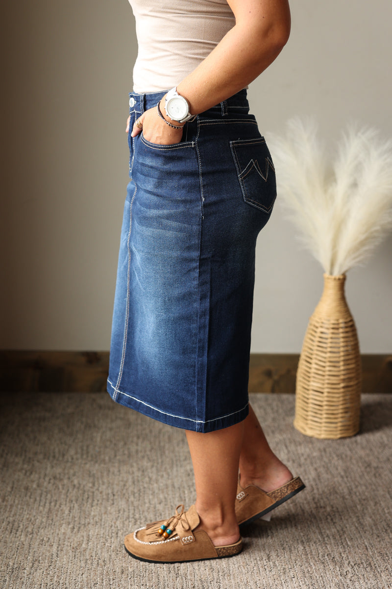 A person wearing a knee-length blue denim skirt with contrast white stitching, paired with tan mule sandals and a light top, standing indoors next to a woven vase with dried pampas grass.