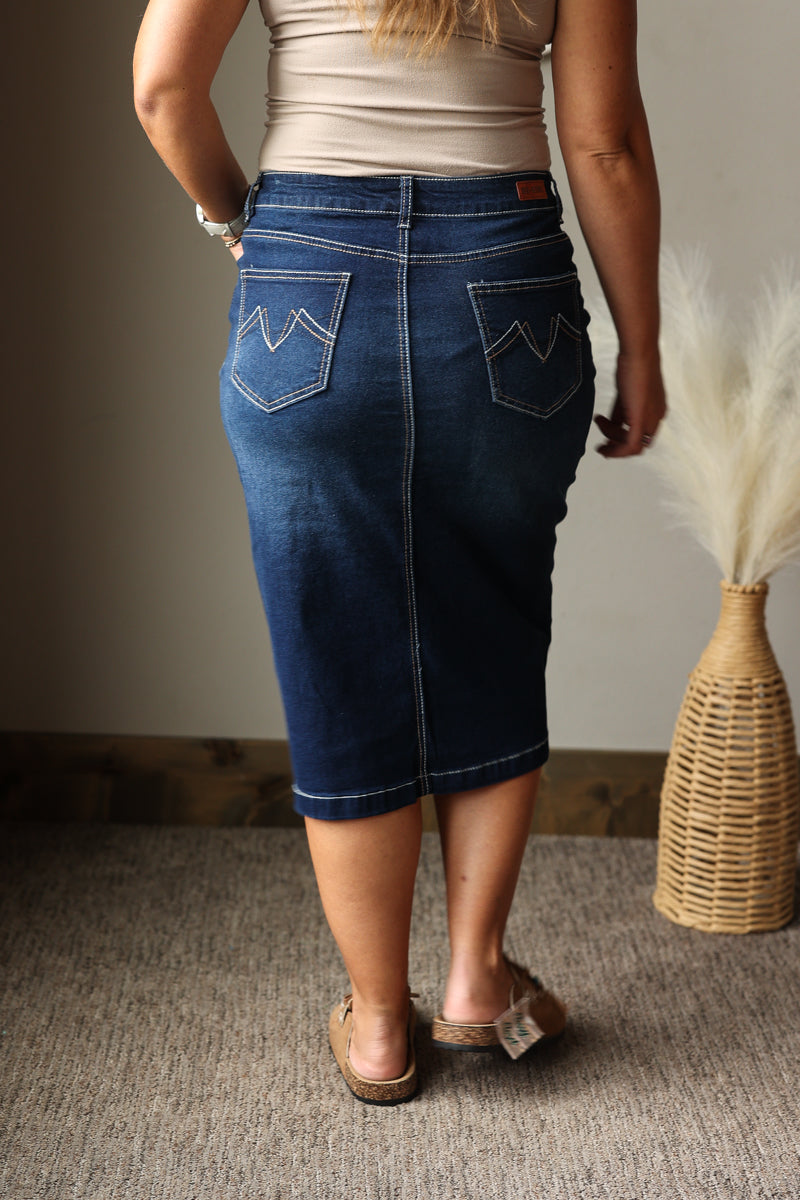 A person shown from the back wearing a dark blue denim pencil skirt with zigzag pocket details, paired with a beige top and brown sandals, standing on a carpeted floor next to a woven vase with dried grass.