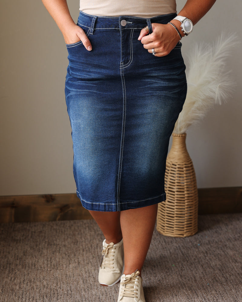 A person wearing a dark blue denim pencil skirt with rolled hems, paired with a beige top and beige sneakers, standing in an indoor setting with a woven vase in the background.