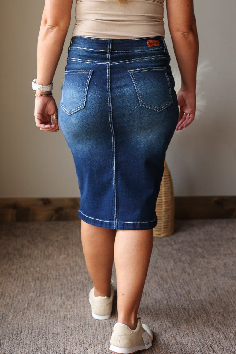A person shown from the back wearing a fitted blue denim pencil skirt with contrast white stitching, paired with a beige top and white sneakers in an indoor setting.