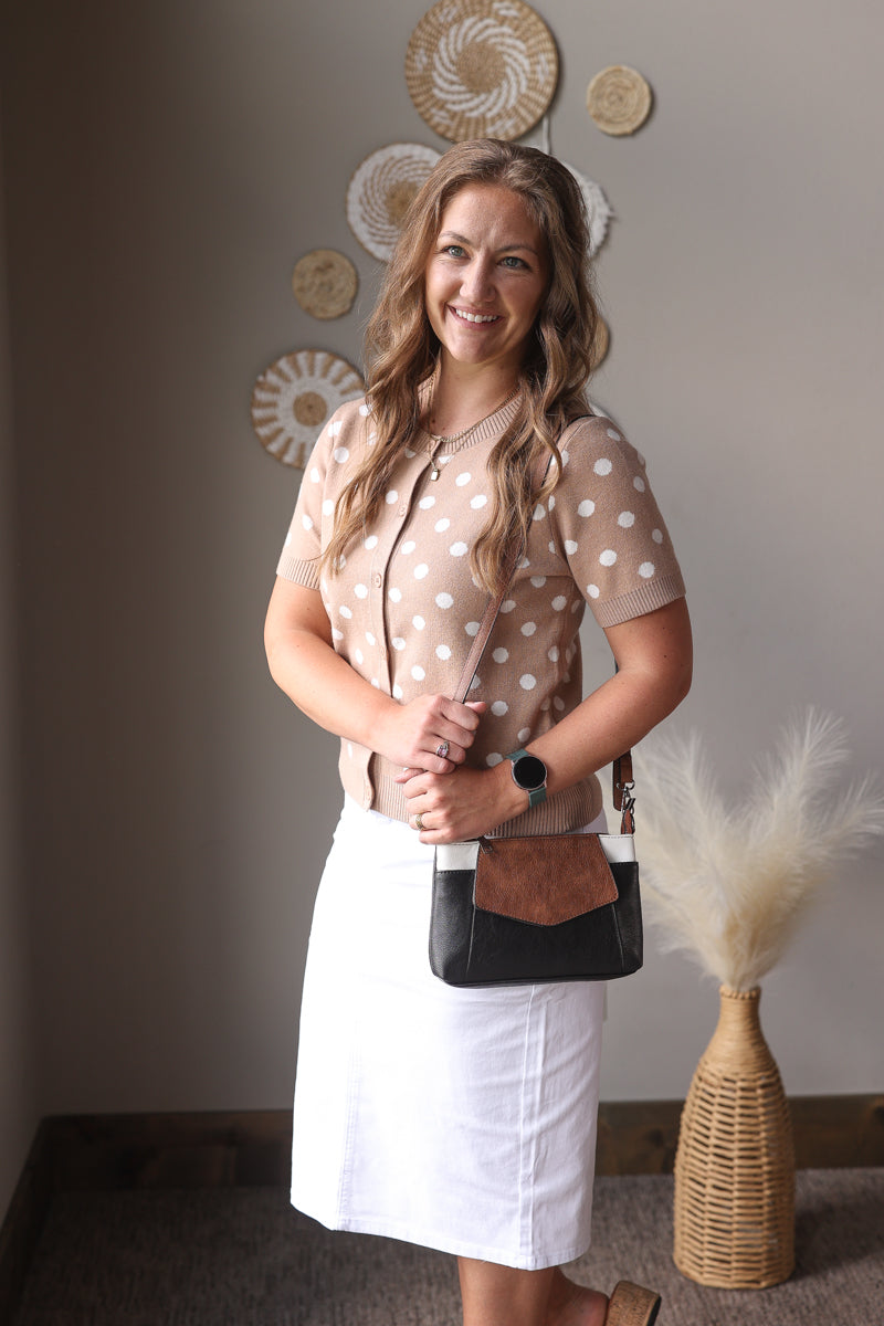 Woman wearing a polka dot blouse and white skirt holding a black handbag in a room with decorative wall art.