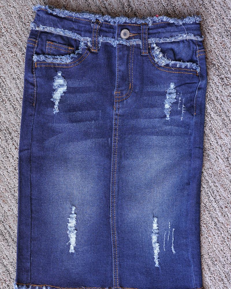 Front flat lay of a girls’ dark wash denim midi skirt with distressed details, front pockets, and a frayed hem.