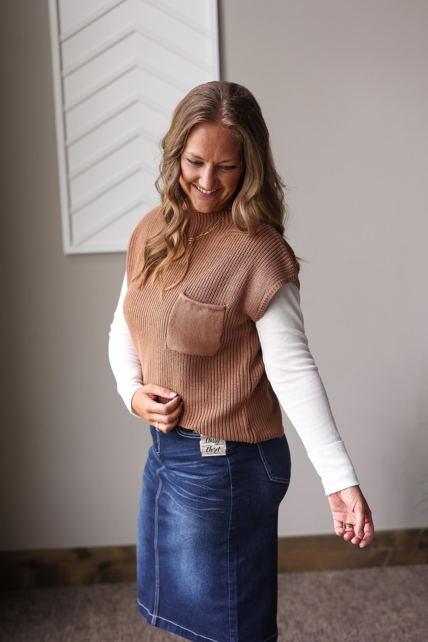 Woman wearing a brown vest over a white long-sleeve shirt and blue denim skirt indoors.