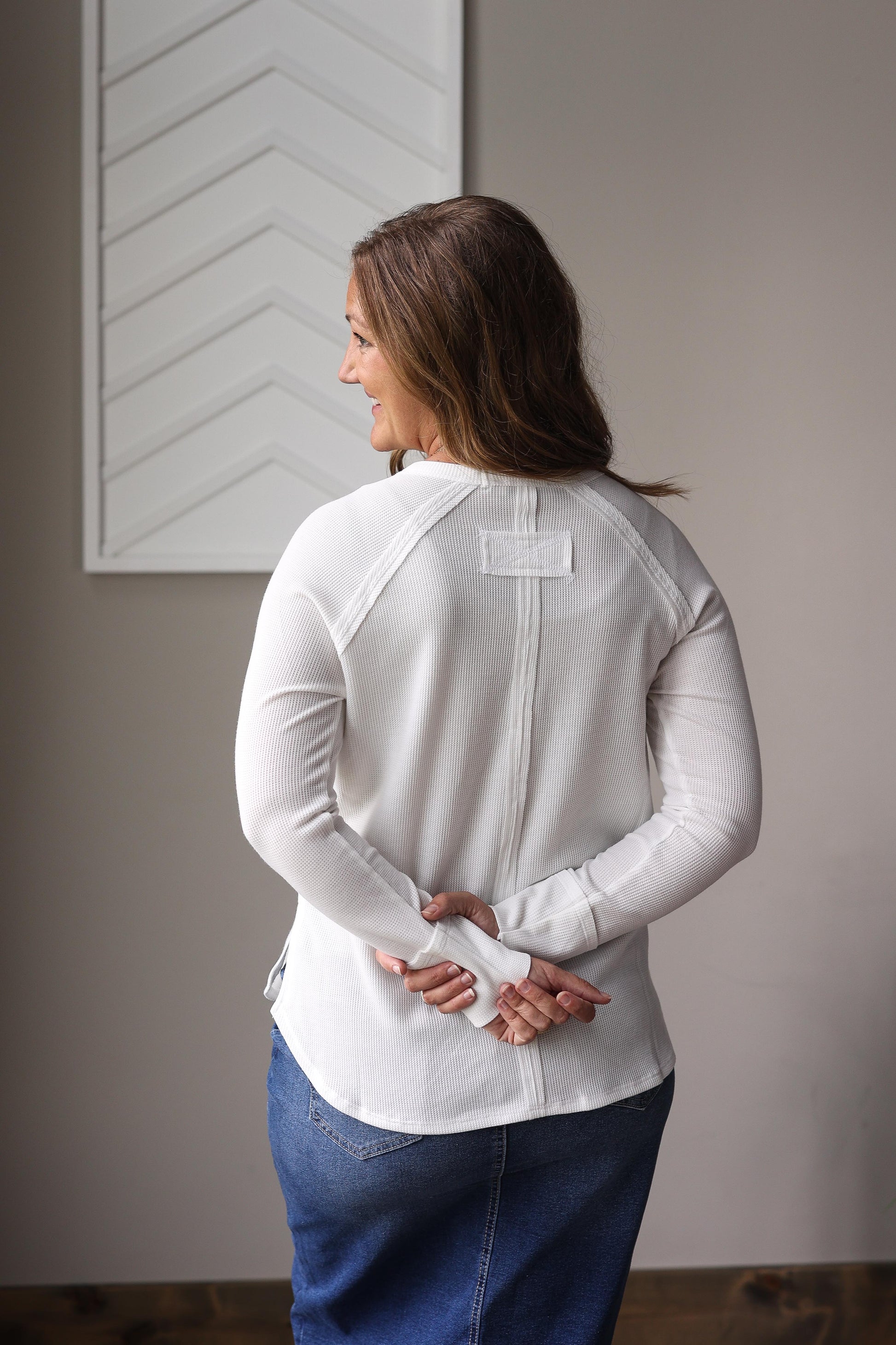 Woman wearing a white long-sleeve shirt with a visible brand logo, standing against a neutral wall.