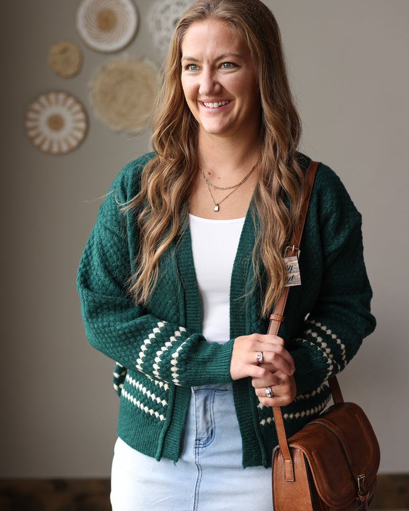 Emerald green textured cardigan with cream diamond stripe cuffs and hem, worn open over a white top and light denim skirt.