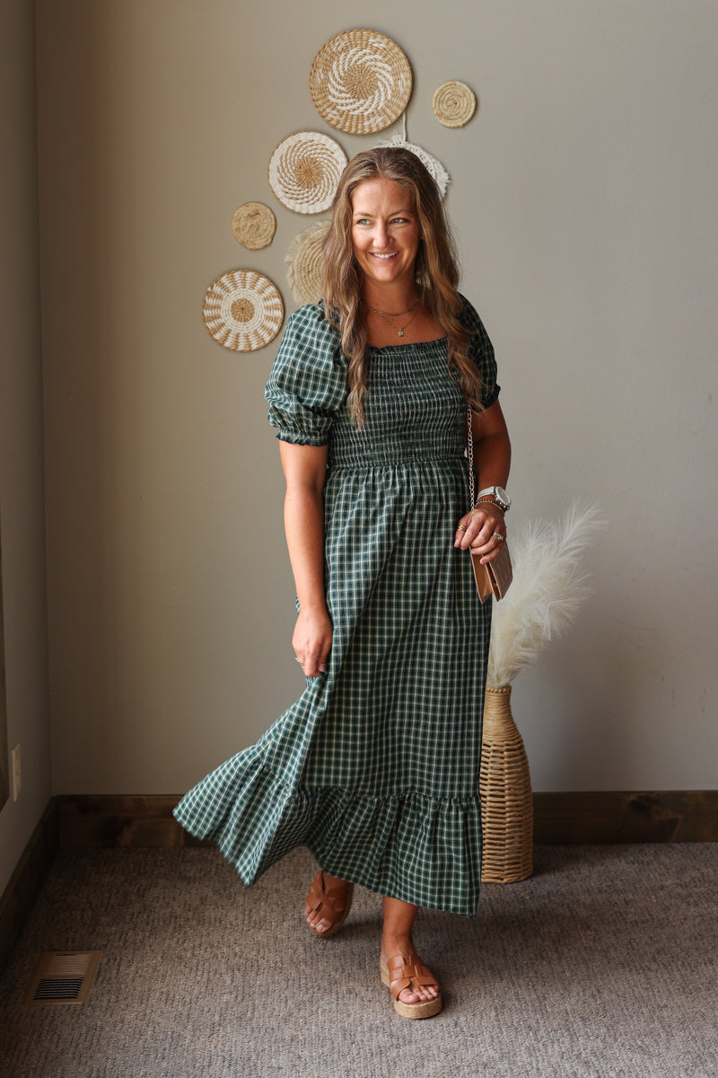 A woman wearing a green and white gingham smocked dress with puff sleeves and a ruffled hem, standing in a room with a beige wall decorated with circular woven wall art.