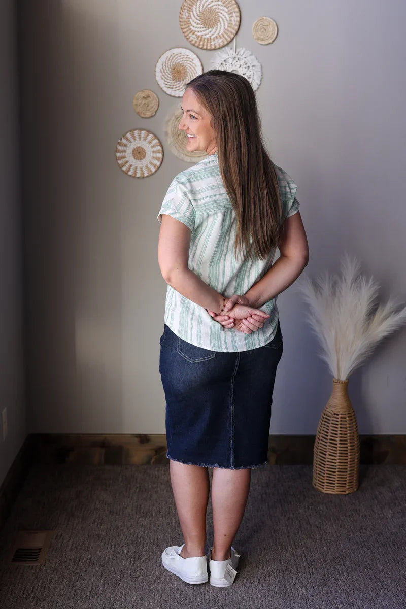 Woman standing in a room with decorative wall items and a vase wearing a green white striped blouse and black denim skirt