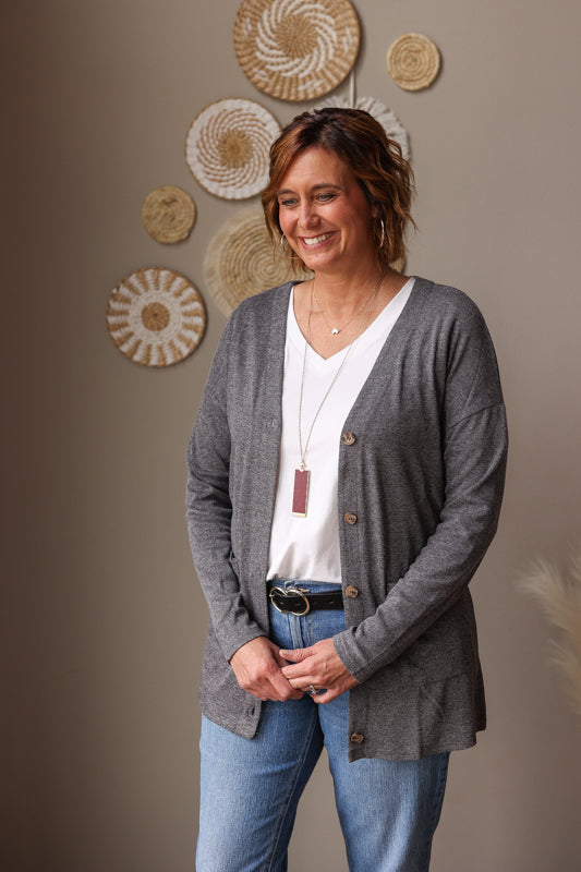 Woman wearing a gray cardigan over a white shirt with a necklace, standing against a wall with decorative baskets. This grey pocketed button front cardigan combines comfort and style, perfect for casual workdays, church, or date nights.