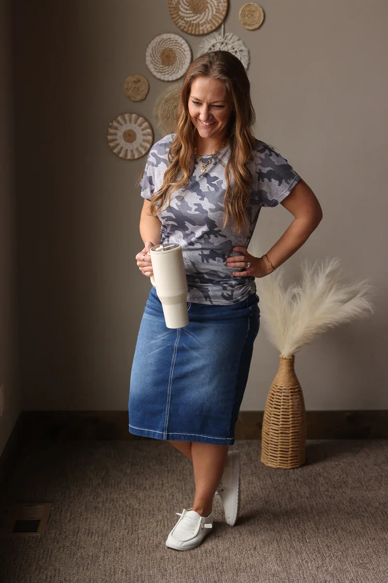 Woman in a room holding a cup, wearing a patterned grey camo shirt and denim skirt.