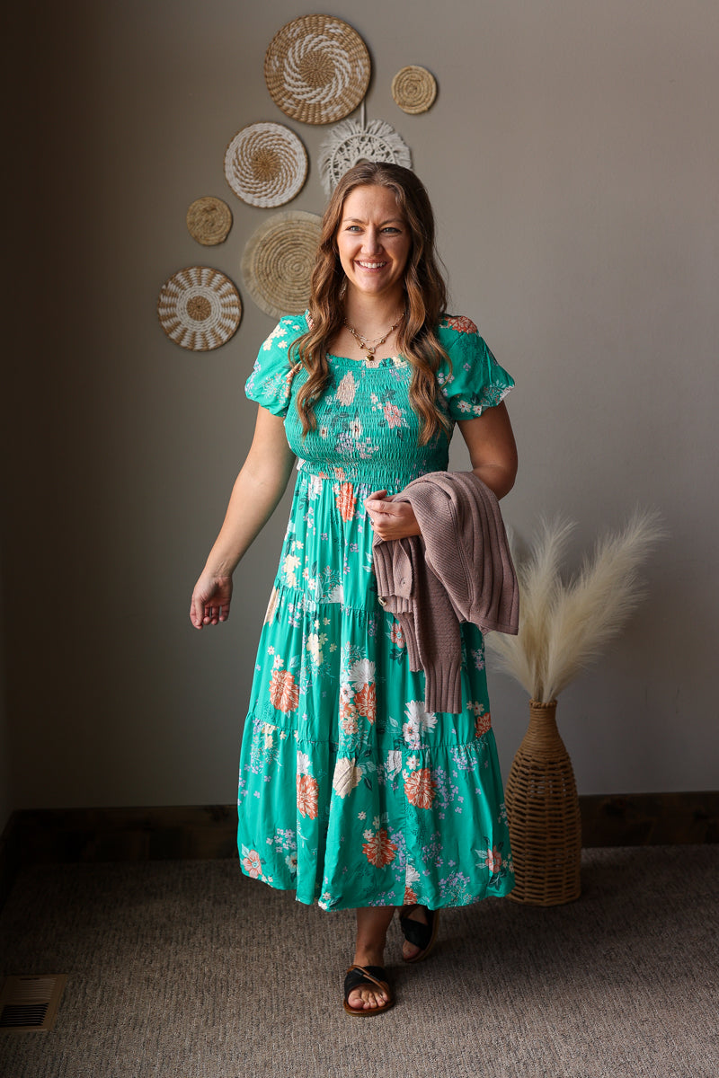 Woman in a teal floral dress standing in a room with decorative wall art and a plant.