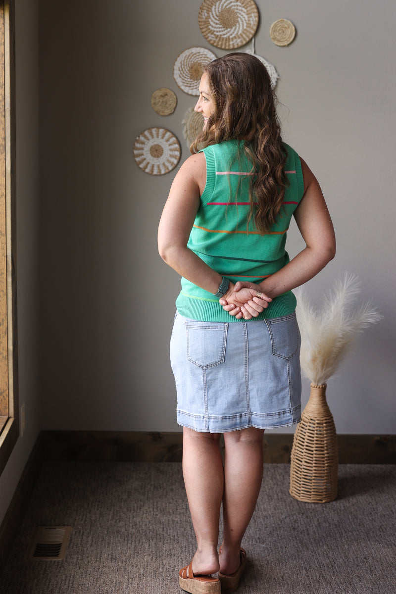 Woman in a green sleeveless top and denim skirt standing in a room with decorative wall art.