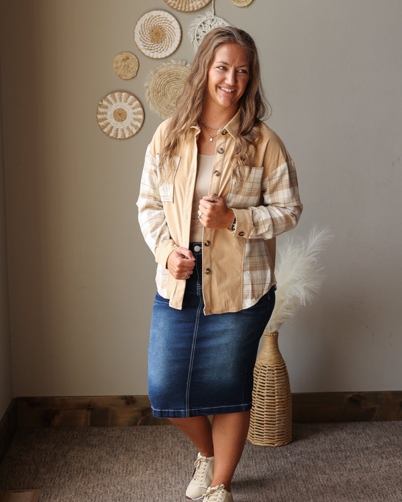 A woman modeling a beige and white plaid button-up shirt jacket over a cream top, paired with a blue denim skirt and beige sneakers, standing in a room with boho wall decor.