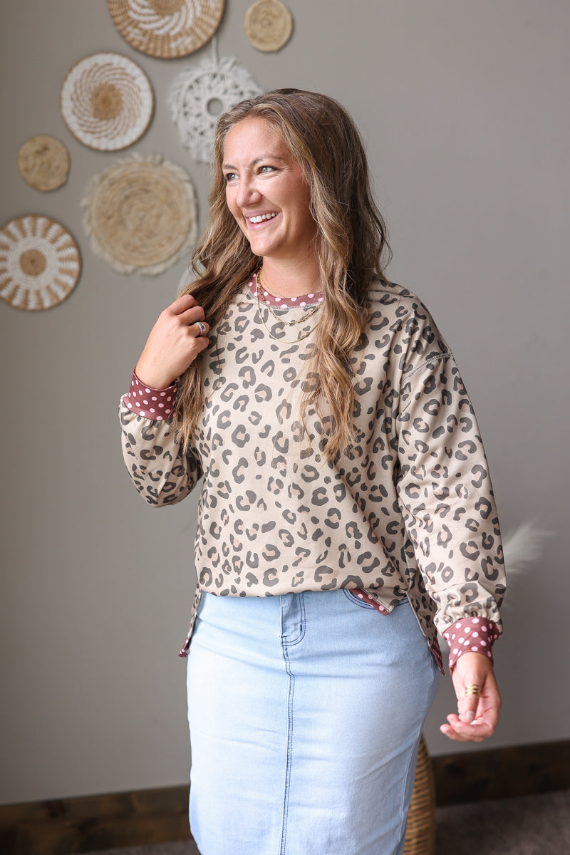 A woman wearing a beige leopard print long-sleeve top with red and white polka dot cuffs, paired with a light blue denim skirt, standing in a room with decorative wall hangings.