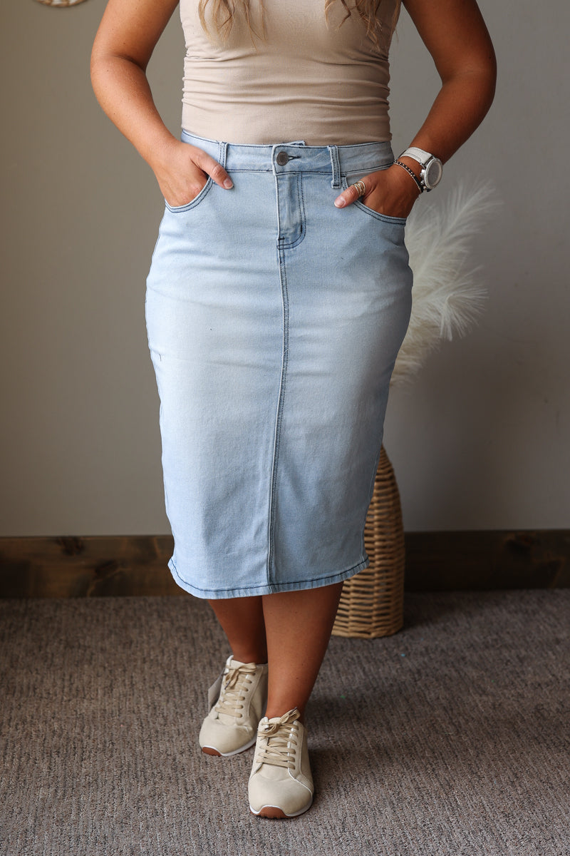A person wearing a light blue denim midi skirt with a front fly and side pockets, paired with a beige top and white sneakers, standing indoors against a neutral wall.