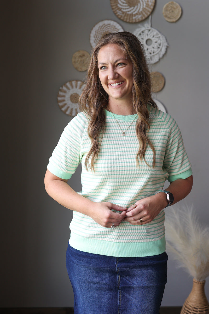 Woman wearing a green and white striped shirt with blue jean skirt, standing against a neutral wall with decorative items.