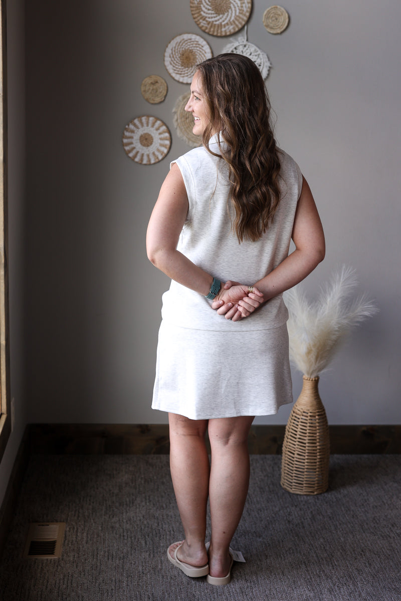 Woman wearing a grey sleeveless top and skirt standing in a room with decorative wall art and a vase.