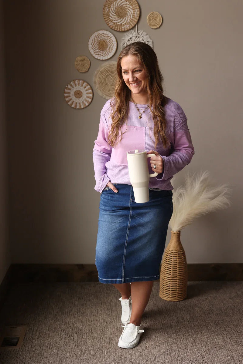 Woman in a purple spring top and blue skirt holding a white mug, standing in a room with decorative wall art.
