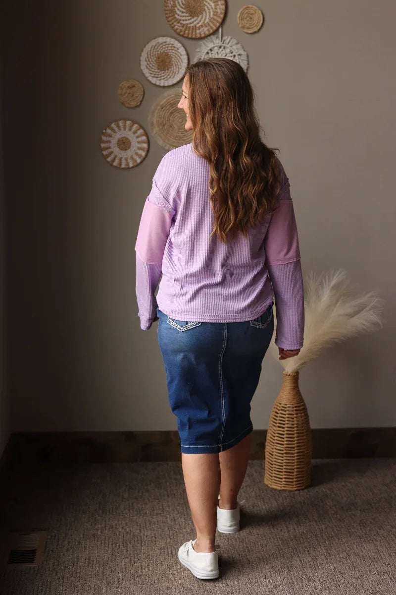 Woman in a purple sweater and blue denim skirt, standing against a wall with decorative plates.