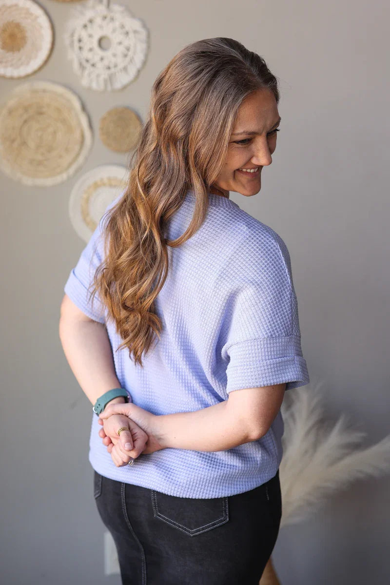 Woman wearing a light purple checkered shirt against a neutral wall with decorative items.