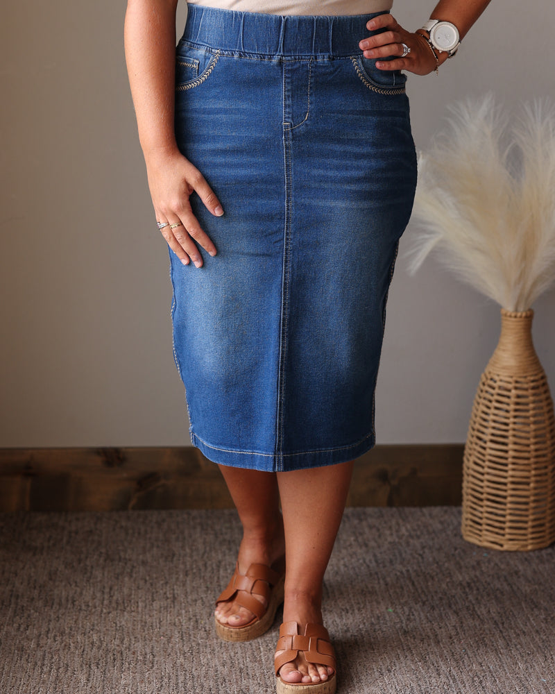 A woman wearing a mid-length blue denim skirt, paired with a beige top and brown platform sandals, standing in an indoor setting with a neutral wall and a decorative woven vase.
