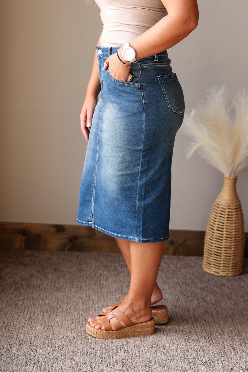 A person wearing a medium-wash blue denim midi skirt with a faded wash, paired with a beige top and brown platform slide sandals, standing on a textured gray carpet.