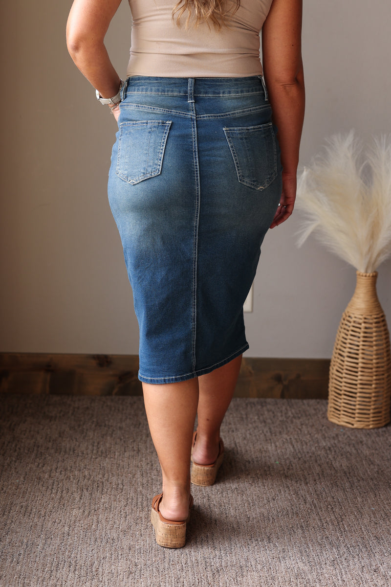 A woman wearing a blue denim midi skirt with back pockets, paired with a beige top and wedge sandals, shown from behind in an indoor setting.
