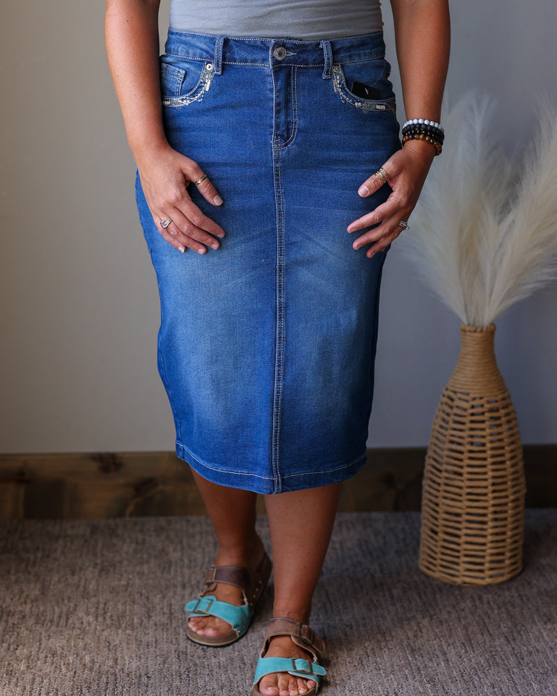 A person wearing a medium-wash blue denim pencil skirt with decorative silver embroidery on the front pockets, paired with a gray top and turquoise sandals, standing indoors next to a woven vase with dried grass.