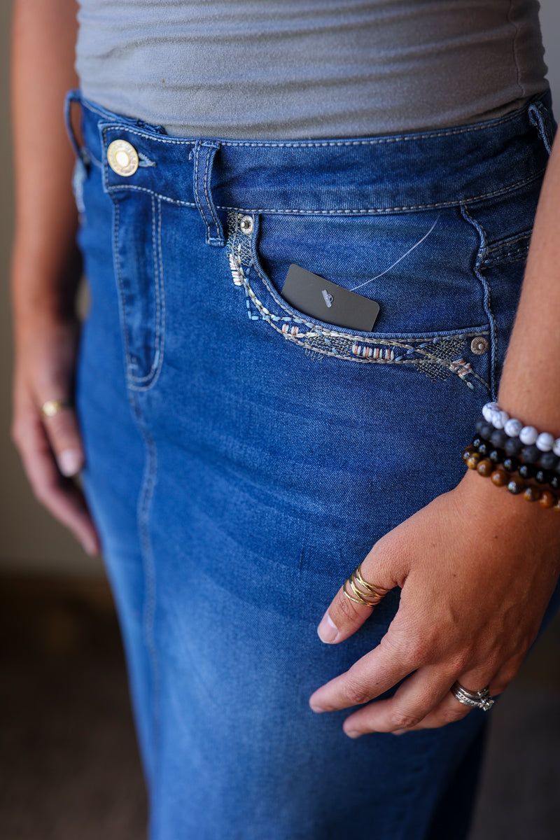 Close-up of a person wearing blue denim jeans with a decorative embroidered pocket and a black product tag tucked into the pocket.