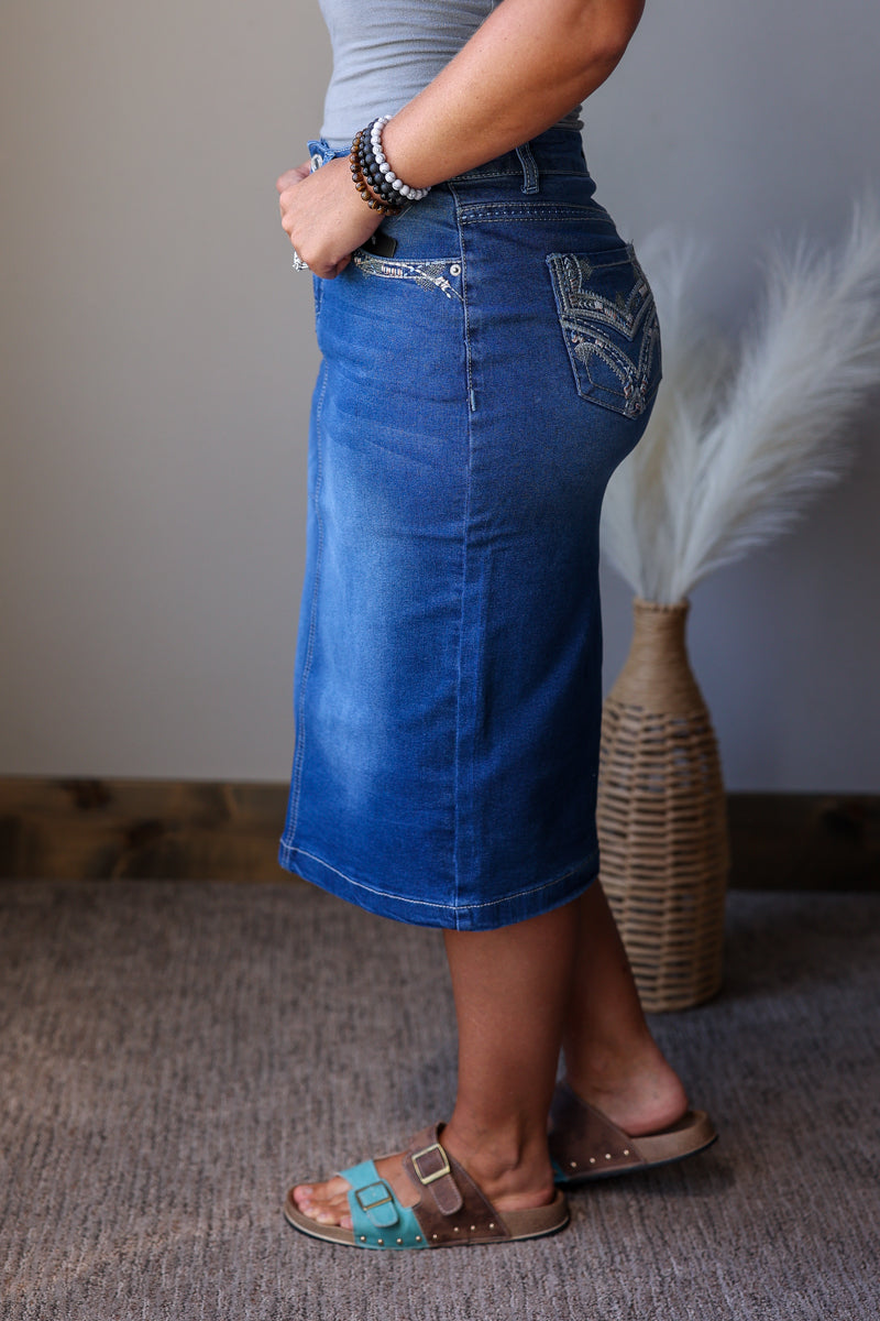A person wearing a medium blue denim midi skirt with decorative silver embroidery on the back pocket, paired with brown slide sandals and beaded bracelets, standing in an indoor setting.