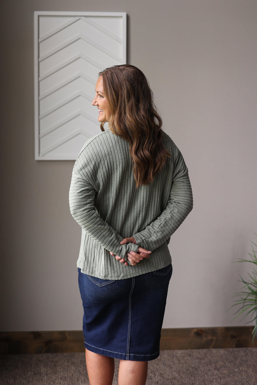Woman wearing a green sweater and blue skirt standing in a room with a neutral wall and plant.