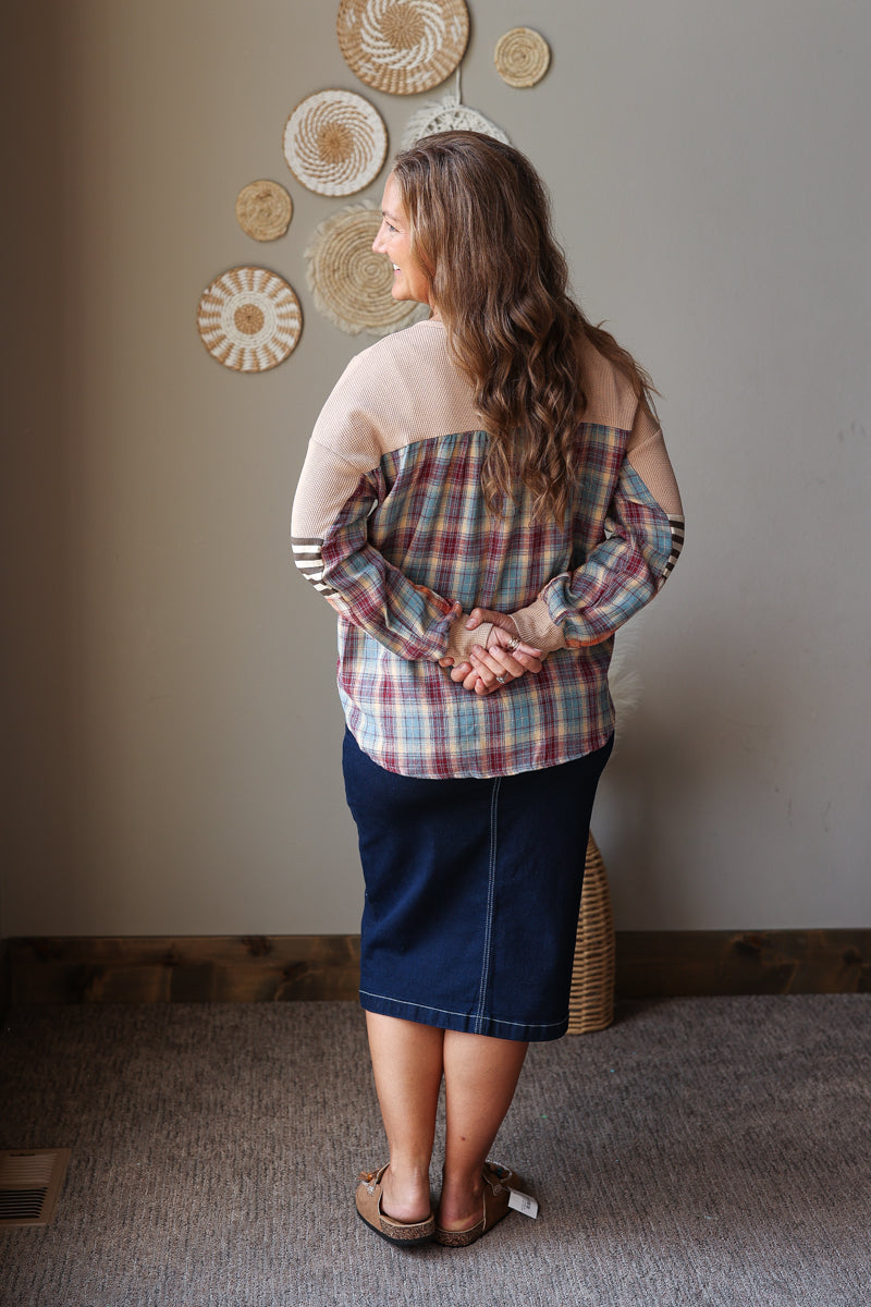 A woman stands with her back to the camera wearing a long-sleeve plaid shirt with beige sleeves and a dark navy blue skirt, standing in a room with a neutral wall decorated with circular wall art.
