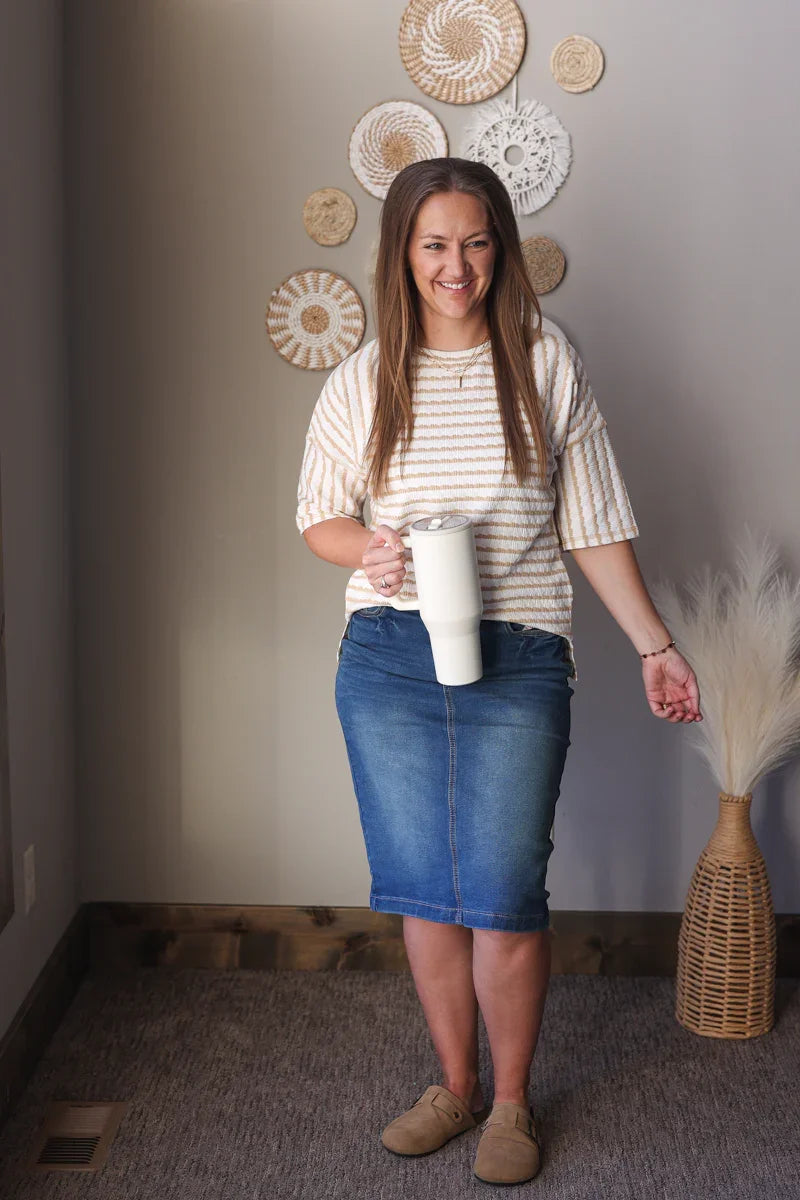 Woman holding a white mug in a room with decorative plates on the wall, wearing a beige white striped half sleeve top for casual spring office outfits