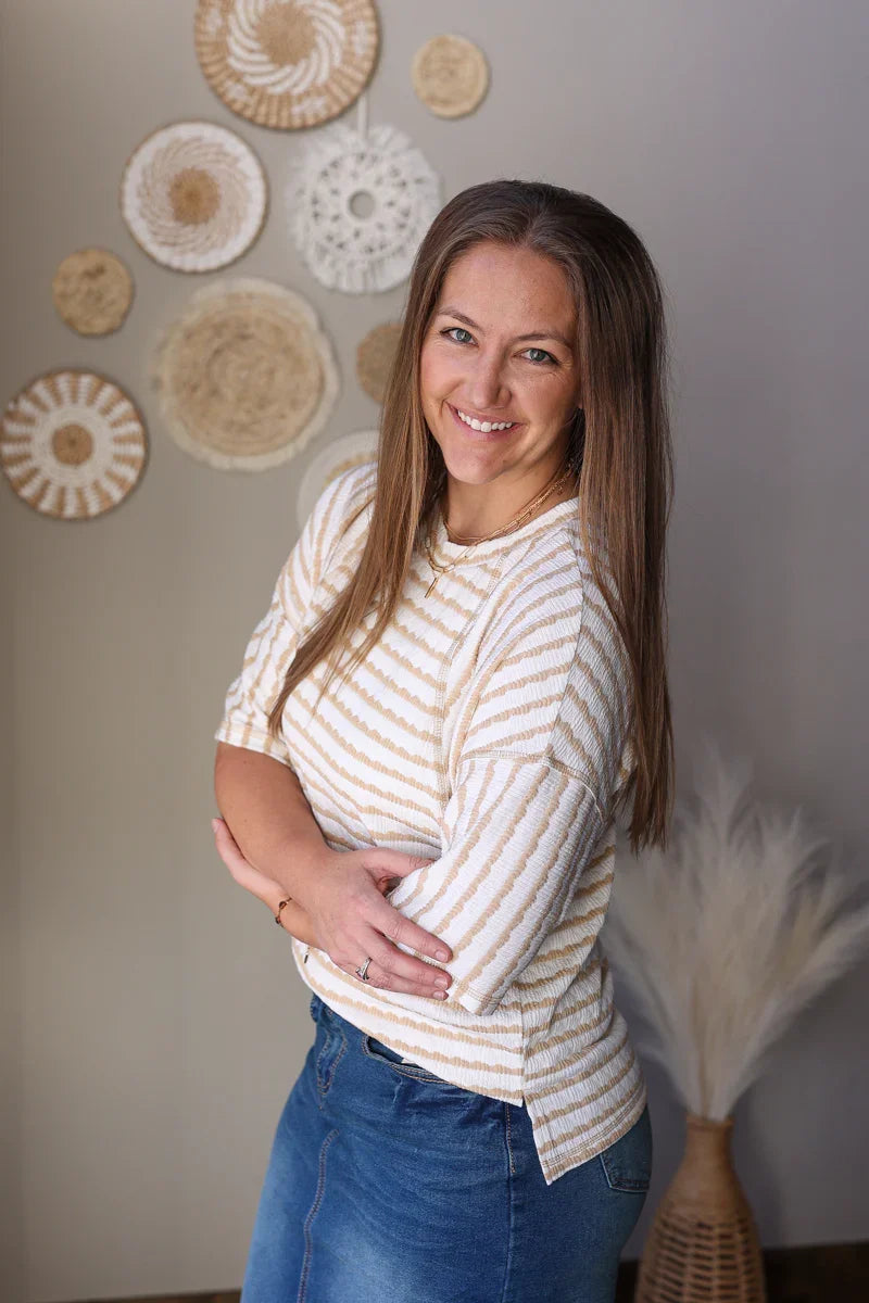 Woman wearing a striped shirt and blue jean skirt standing in front of decorative wall art.