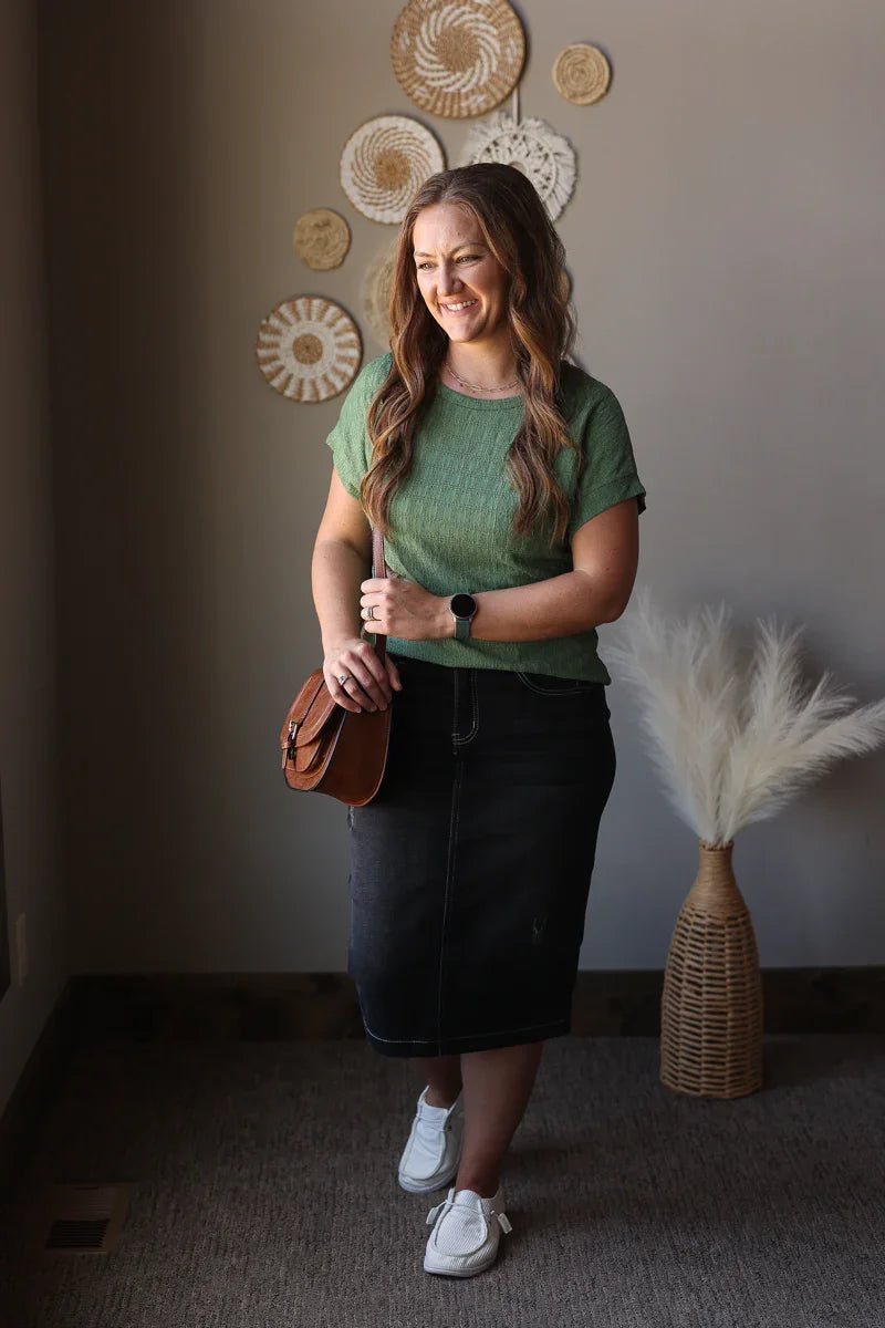 Woman in green top and black skirt standing indoors with decorative elements on the wall.