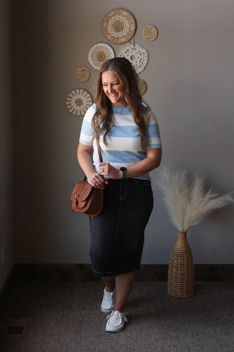 Woman in a striped shirt and skirt standing in a room with decorative wall art and a vase.
