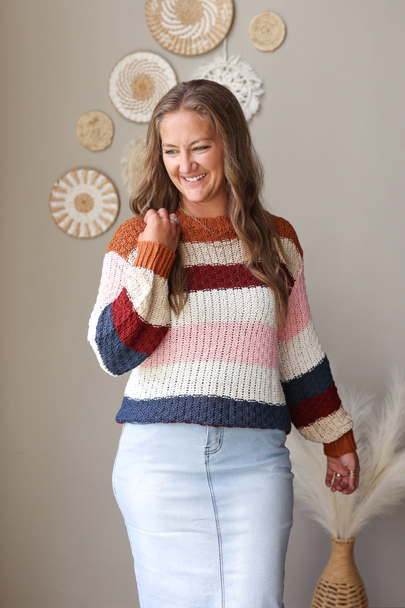 A woman wearing a chunky knit striped sweater with horizontal bands of cream, rust orange, burgundy, light pink, and navy blue, paired with light-wash blue jean skirt. She is standing in a room with a beige wall decorated with circular woven wall art and a vase with dried grass.