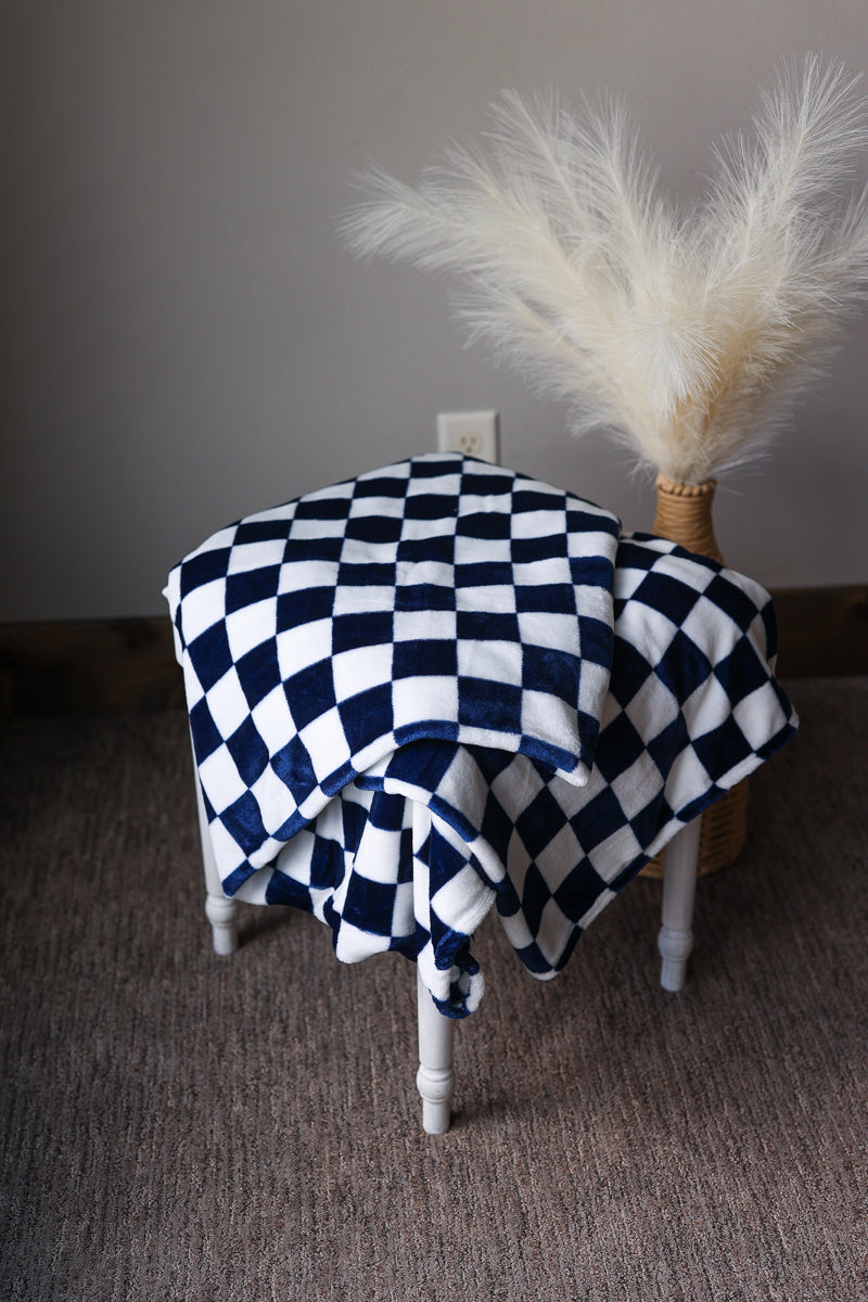 Black and white checkered blanket draped over a chair with a decorative vase of pampas grass on a plain background