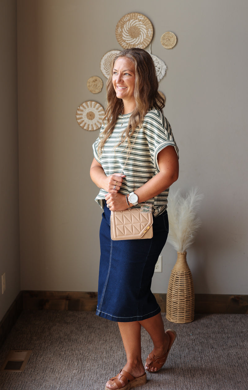 A woman posing in a home interior wearing a green and white striped t-shirt, a dark blue denim midi skirt, and brown leather sandals, while holding a beige quilted crossbody bag.