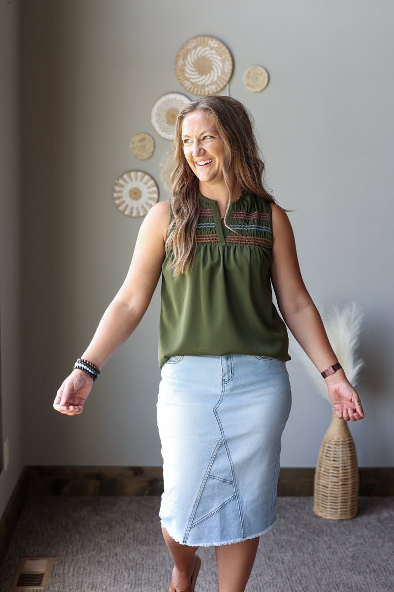 A woman modeling a sleeveless olive green top with colorful embroidered detailing at the neckline and a light blue denim skirt, standing in a room with boho-style wall decor.