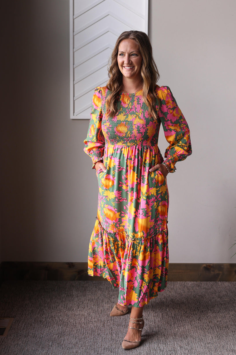 Woman wearing a colorful floral dress standing indoors against a neutral wall.
