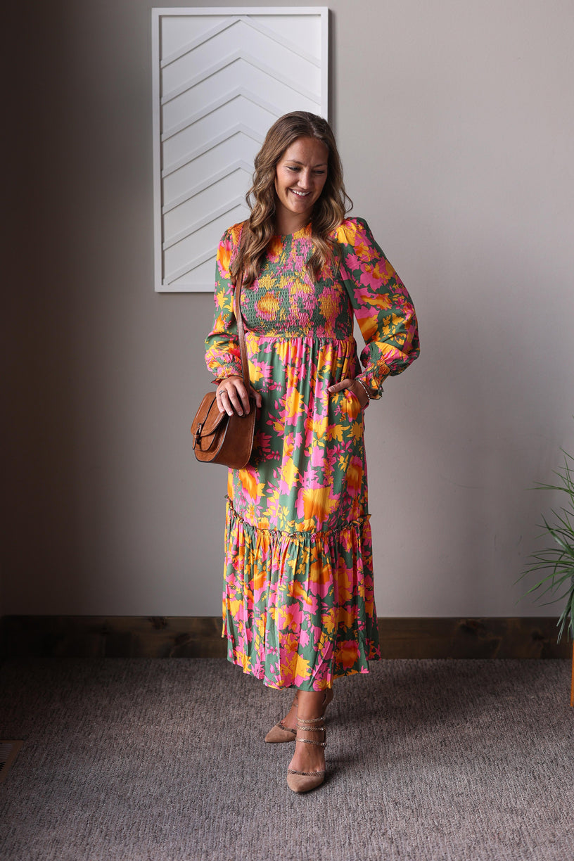 Woman wearing a colorful floral dress standing indoors against a plain wall.