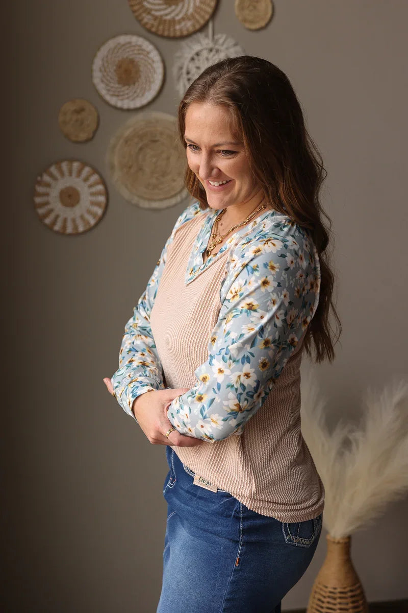 Woman wearing a floral blouse over a pink top with blue jeans skirt, standing against a wall with decorative circular patterns.