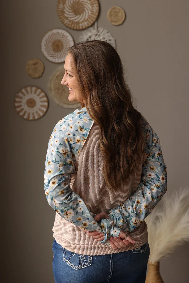 Woman wearing a floral long-sleeve shirt with a neutral sweater underneath, standing against a wall with decorative plates.